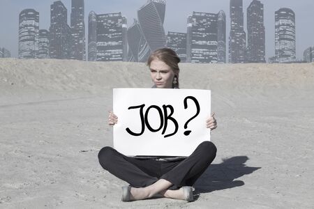 young woman is looking for a job with inscription in hands sitting on the sand in desert. sitting on the sand in the desert. Skyscrapers of the modern city in backgroundの写真素材