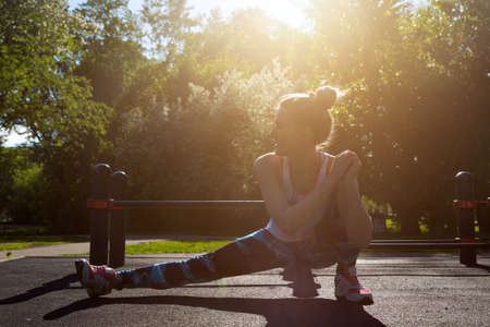young blonde girl is sitting in a muscle stretching pose, warming up before training in a city park, a sunny bright day.の写真素材