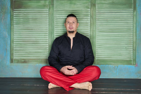 Portrait of a young man, black shirt and red slacks, hairstyle with shaved temples and slicked- back hair at the top of the head, different emotions, green wall, brown floor, beard, lotus postureの写真素材