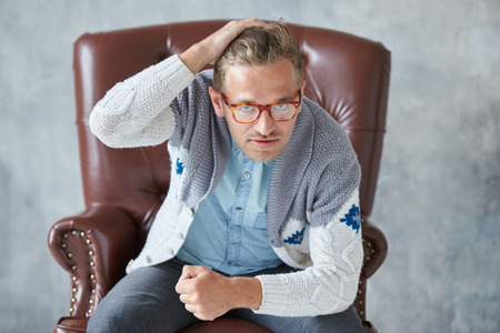 Portrait of a stylish intelligent man with glasses stares into the camera, good view, small unshaven, charismatic, blue shirt, gray sweater, sitting on a brown leather chairの写真素材