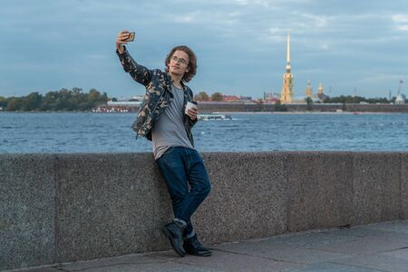 The handsome men is waiting someone, drinks coffee and looks in phone, he is dressed in a military jacket and jeans, Peter and Paul fortress is on backgroundの写真素材