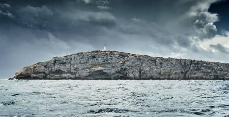 Lighthouse is on edge of the rock, the landscape of the balearic sea and improbable mountains, azure water, the storm sky, mountainsの写真素材
