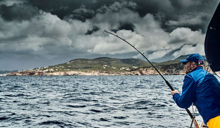 The fisherman fishes from the boat, he is dressed in blue jacket and cap, the landscape of the balearic sea and improbable mountains, azure water, the storm skyのeditorial素材