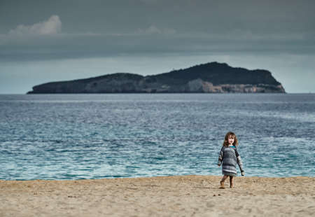 The little cute girl is on a beach, long curly hair, she is dressed in sweater, The Landscape of the balearic, azure water, cloudy sky, lonely mountain is on backgroundのeditorial素材