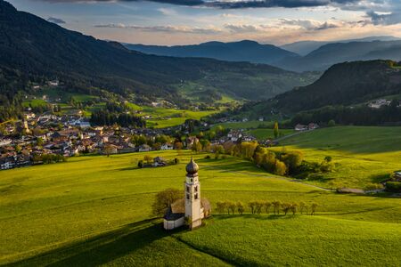 Aerial view of picturesque valley with chapel in sunset, Trentino, amazing green meadows of the mountains of Italy, roof tops of houses, Dolomites on background, drone flies around chapelの写真素材