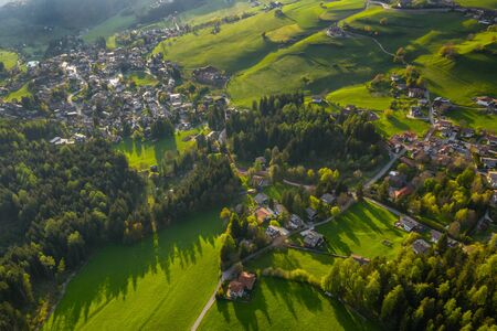 Aerial view of improbable green meadows of Italian Alps, green slopes of the mountains, huge clouds over a valley, roof tops of houses, Dolomites on background, sunshines through cloudsの写真素材