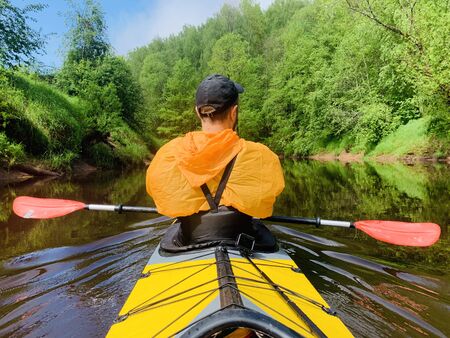 The men in a cap and raincoat of orange color floats on a kayak on the forest quiet river, the beautiful landscape, a changeable weather, actively rows with an oar, beautiful reflectionの写真素材