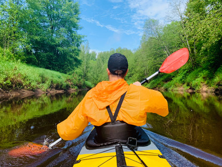 The men in a cap and raincoat of orange color floats on a kayak on the forest quiet river, the...