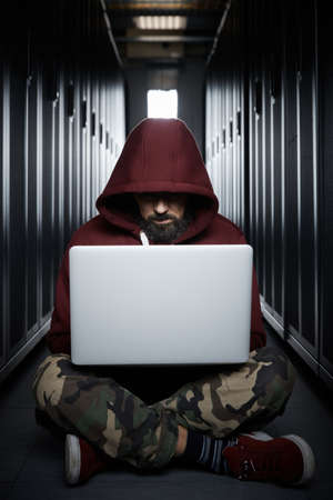 The hacker in a hood sits with the computer among servers, the burglar, face covered with a hood. Storage of data, cloudy services, system breaking, attack of hackersの写真素材