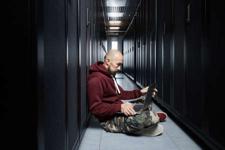 The hacker with a bald head and a beard sits with the computer among servers, the burglar, Storage of data, cloudy services, system breaking, attack of hackersの写真素材