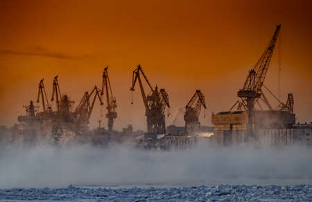 The construction of nuclear icebreakers at magic sunset, cranes of of the Baltic shipyard in a frosty winter day, steam over the Neva river, smooth surface of the river, sky of orange colorの写真素材