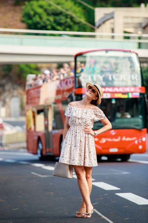 a bright beautiful girl in a light dress and hat walks along the streets of Monaco in sunny weather in the summer, tourist red bus in the backgroundの写真素材