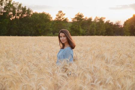 The beautiful young girl with long brunette hairs poses in the field with wheat at sunset, she...