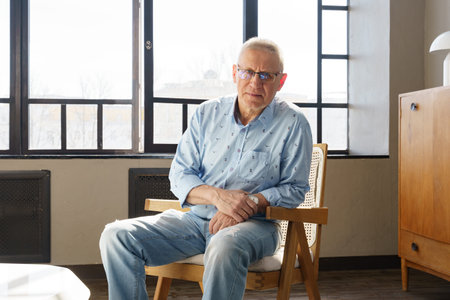 An elderly man in glasses with diopters is siting on the chair in the living room, he look into the...