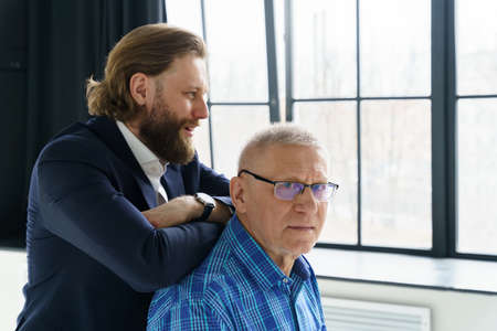 the son in a stylish suit leans on the shoulders of his father, Father and son in huge window background, both men look into the camera, an elderly man in glasses, young man with a beard and long hairの写真素材