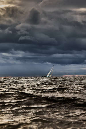 Sailboat in sea at stormy weather, blurred background, stormy clouds sky, sail regatta, reflection of sail in the water, big waves of waterの写真素材