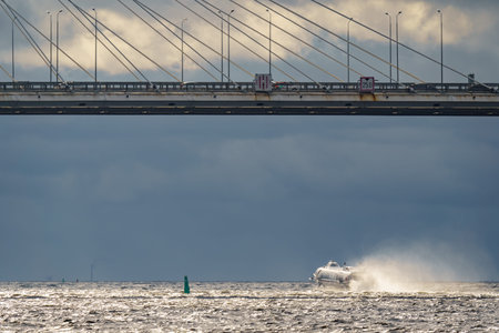 a high-speed passenger hydrofoil boat passes under a cable-stayed bridge in sunny weather, dark...