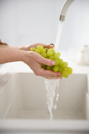 Close up view of a woman washes fresh green grapes under the pressure of flowing water in the white kitchenの写真素材