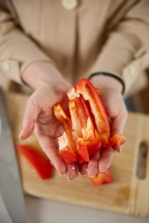 A womans hands hold pieces of sliced red sweet pepper, top view, a wooden chopping board and a...