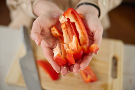 A womans hands hold pieces of sliced red sweet pepper, top view, a wooden chopping board and a kitchen knife lie in the backgroundの写真素材