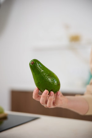 A large avocado in a womans hand in the interiors of a bright and spacious kitchen