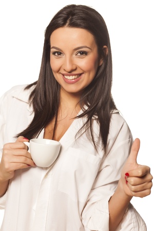 smiling beautiful brunette in a white men s shirt drinking coffee and showing his thumbの写真素材