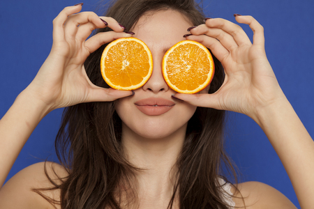 Smiing young woman posing with slices of oranges on her face on blue backgroundの写真素材