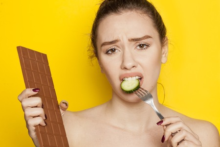 A young beautiful woman eating cucumber and looking in chocolate on yellow baclgroundの写真素材