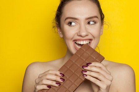 young happy woman enjoying eating chocolate on a yellow backgroundの写真素材