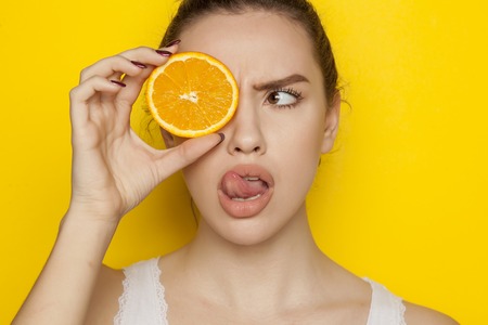 Surprised young woman posing with slice of orange on her face on yellow backgroundの写真素材