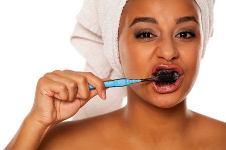 portrait of a happy young dark-skinned woman brushing her teeth with black toothpaste on a white...