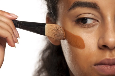 portrait of a young dark-skinned woman applying liquid makeup base with brush on her face on a white backgroundの写真素材
