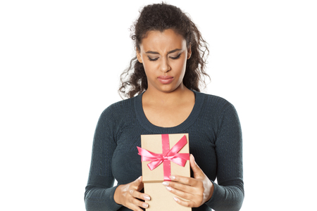 unhappy african woman at the desk with a laptop opens a box with a giftの写真素材