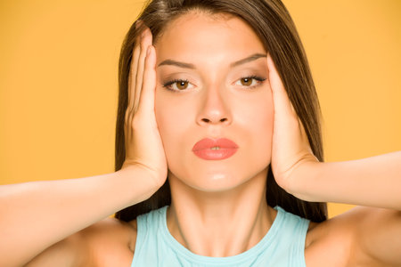a young woman tightens her face with her hands on a yellow background