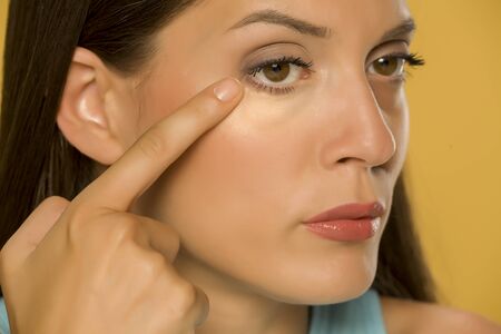 Young woman touching her low eyelids on yellow backgroundの写真素材