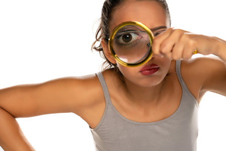 a suspicious young woman looks through a magnifying glass on white background