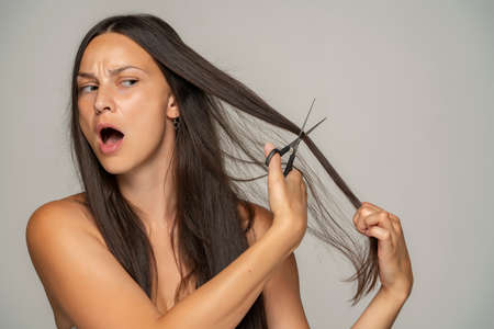 a nervous woman cuts her hair with scissors on a gray backgroundの写真素材