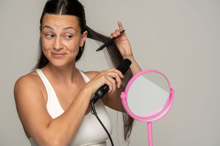 Young smiling woman straightening her hair with a straightener on gray background