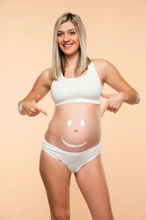 Future mother showing a smiley face drawing on belly on a beige backgroundの写真素材