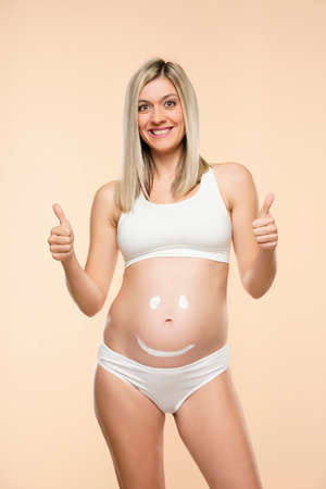 Future mother posing with a smiley face drawing on her belly and showing thumb up on a beige backgroundの写真素材
