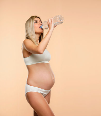 Young pregnent woman in lingerie drinks water from a plastic bottle on a beige backgroundの写真素材