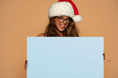 christmas, x-mas, people, advertisement, sale concept - happy woman in blue dress and santa helper hat with blank blue board on a beige backgroundの写真素材