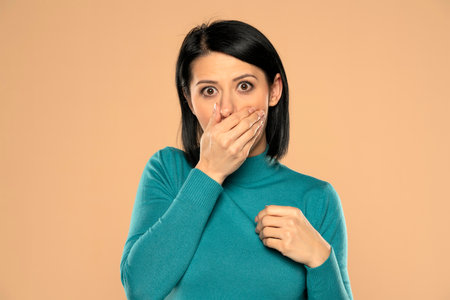 Middle aged woman covers her mouth with her hand on a beige backgroundの写真素材