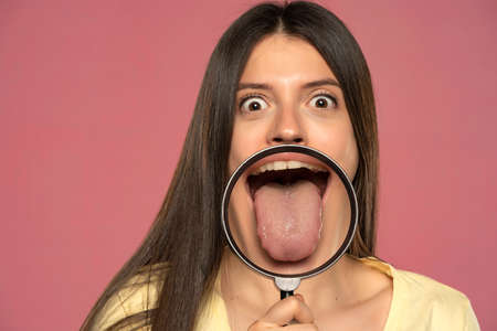 Young happy woman showing her healthy tongue through a magnifying glass on a pink backgroundの写真素材