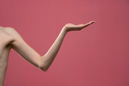 Young woman's stretched skinny arm and open palm holding imaginary product. Isolated on pink background.の写真素材
