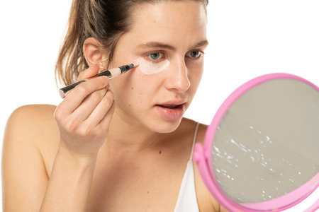 Young woman applyes concealer under her eyes on white studio background.