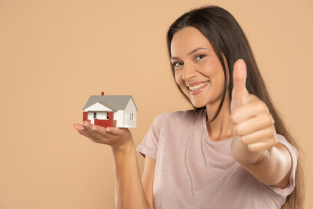 Young woman smiling and holding house sample model isolated over beige studio background, Real estate and home insurance conceptの写真素材