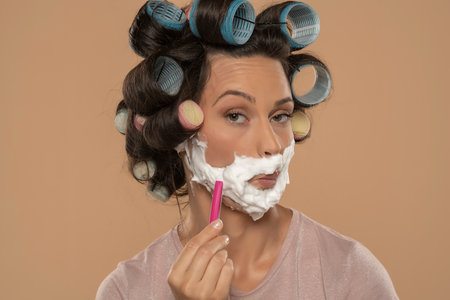 attractive young woman with hair curlers rollers shaves her face on a beige studio backgroundの写真素材