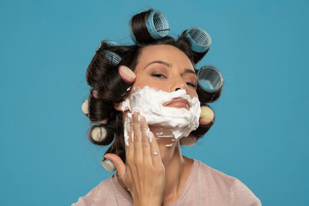 attractive young woman with hair curlers rollers applyes shaving foam on her face on a blue studio backgroundの写真素材