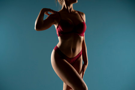 Tanned woman in top form, perfect body shape in the shadow. Parts of woman body in red underwear, front view, blue studio backgroundの写真素材